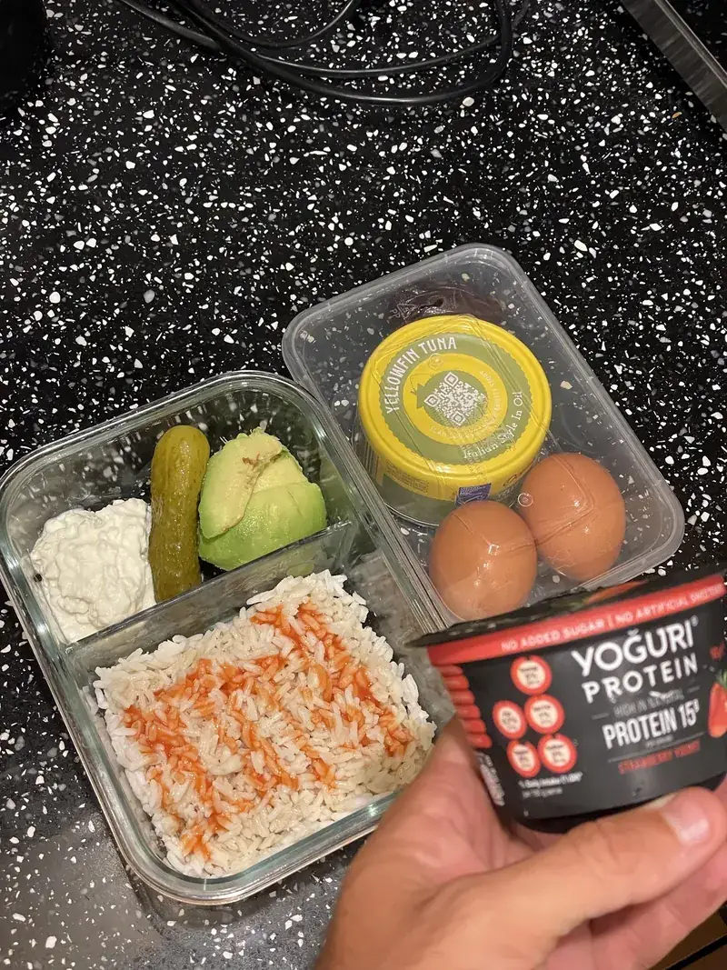 Meal prep containers with rice and hot sauce, cottage cheese, pickle, avocado, a tin of tuna, boiled eggs and a protein yoghurt